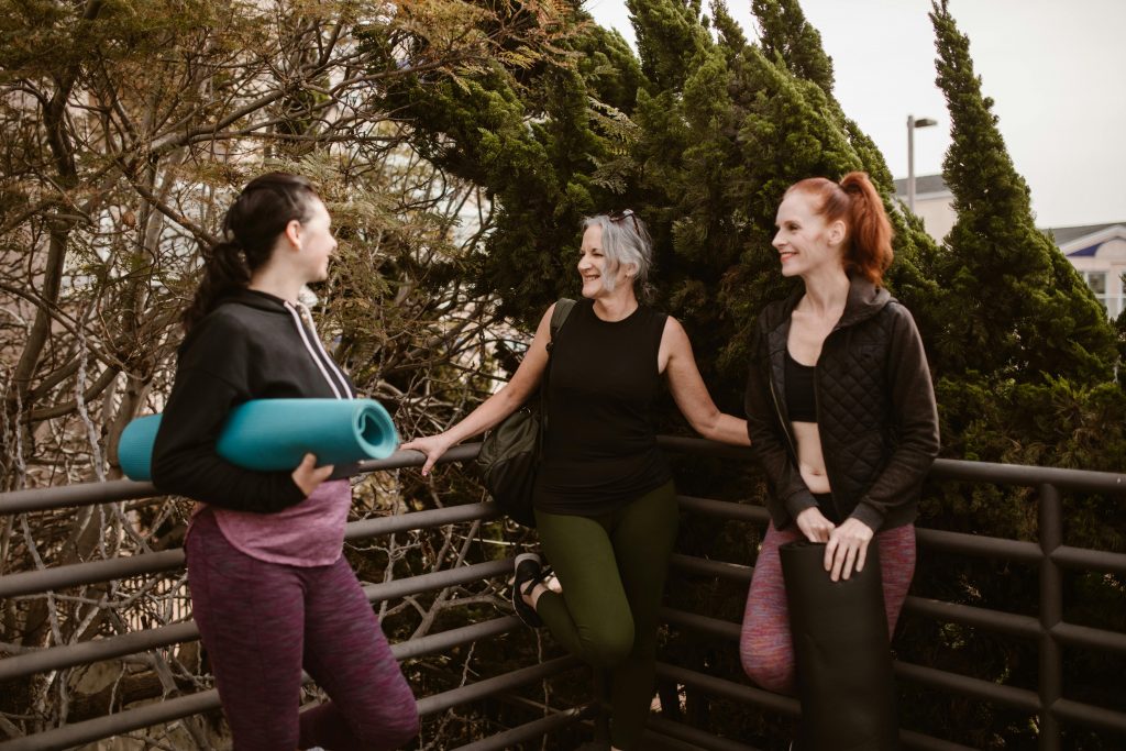 Three women with yoga mats enjoy conversation outdoors, promoting health and wellness.