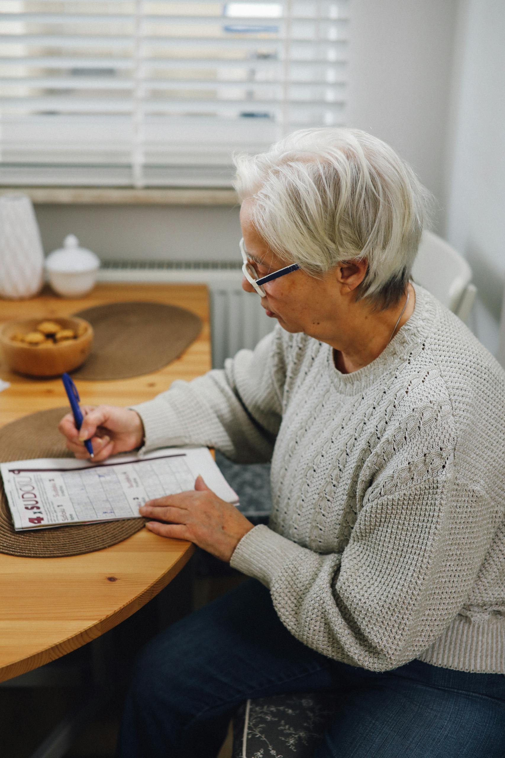 Elderly woman with gray hair writing on paper at a wooden table indoors.