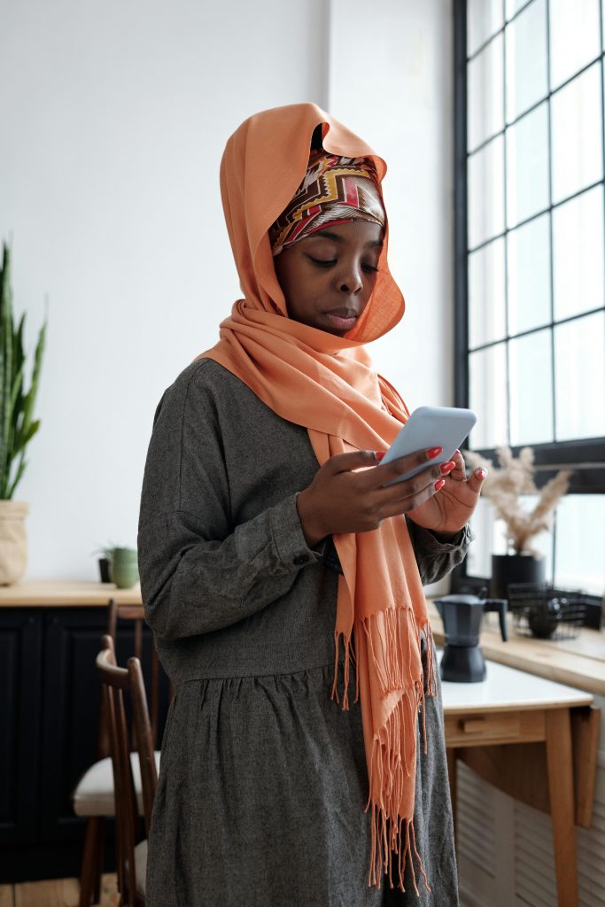 A woman wearing a hijab and headscarf texts on her smartphone indoors, near a large window.