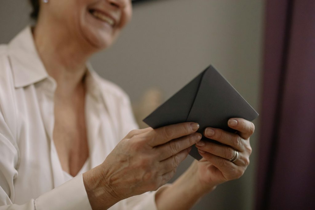 A close-up of a smiling senior woman holding an envelope indoors on a sunny day.