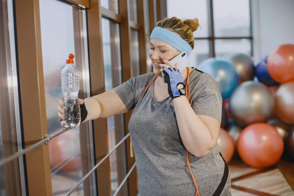 Plus-size woman enjoying a workout session, talking on phone, and staying hydrated in a gym environment.