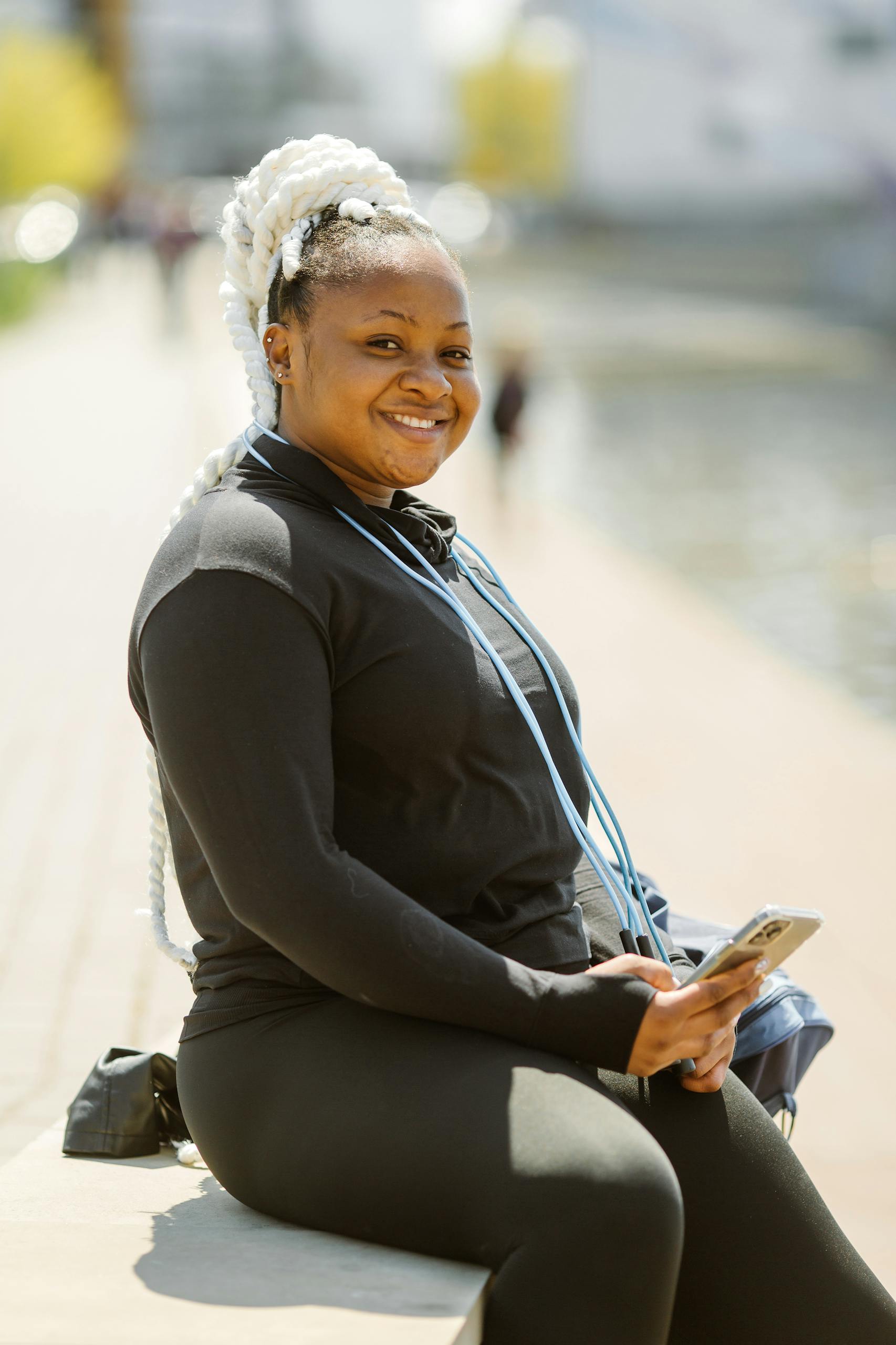 Confident black woman in active wear sitting by the water, smiling while holding her smartphone.