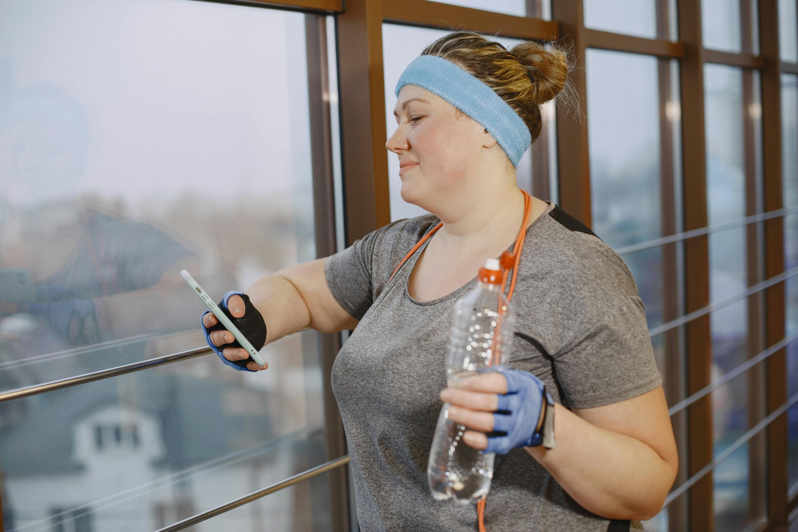 Plus-size woman on phone during indoor exercise, holding water bottle in gym setting.