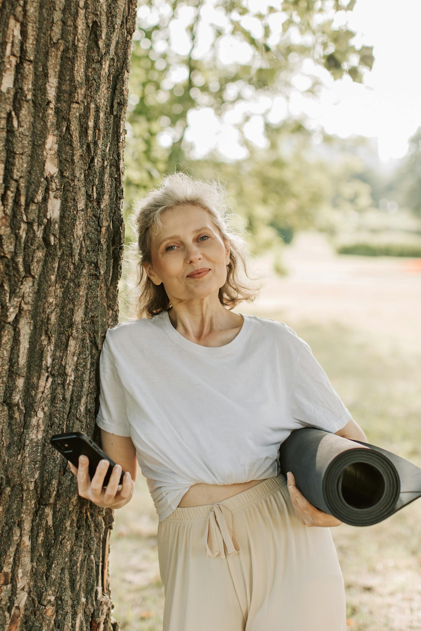 Senior woman leaning on tree with yoga mat and phone, enjoying a sunny day outdoors.
